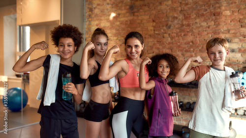 We are strong. Happy female trainer and positive children showing muscles while smiling at camera, standing in gym