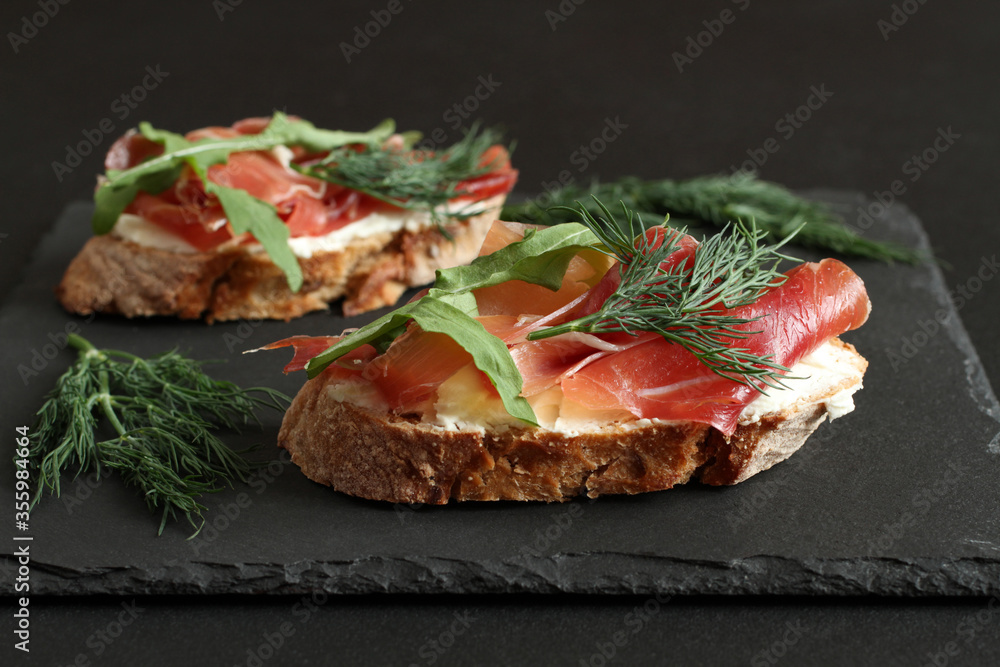 Buckwheat toasts with goat cheese, prosciutto and arugula lying on a black stone board on a black table. Closeup