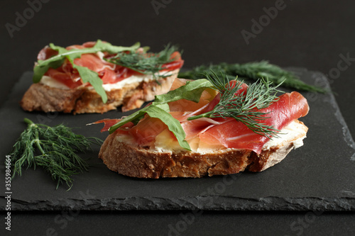 Buckwheat toasts with goat cheese, prosciutto and arugula lying on a black stone board on a black table. Closeup