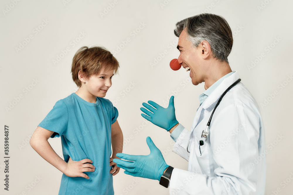 Having fun. Cheerful male pediatrician with clown nose working with happy little boy, standing against grey background Having fun. Cheerful male pediatrician with clown nose working with happy little boy, standing against grey background