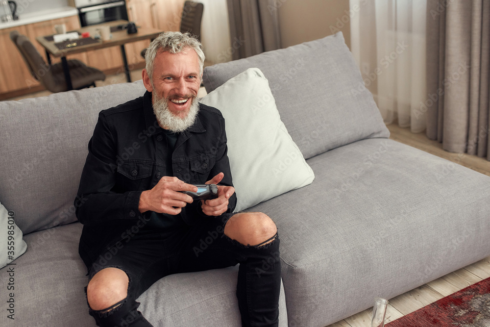 Stay on top. Bearded middle-aged man holding controller, playing video games after smoking marijuana from a bong or glass water pipe sitting on the couch at home