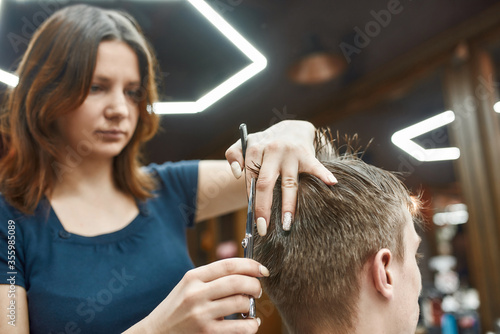 Concentrated at work. Focused barber girl holding scissors and comb in hands and making haircut for her client. Young guy visiting barbershop