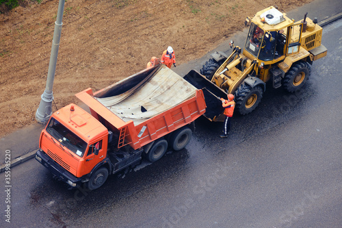 A worker with a shovel pours asphalt from a truck into the bucket of an excavator, top view