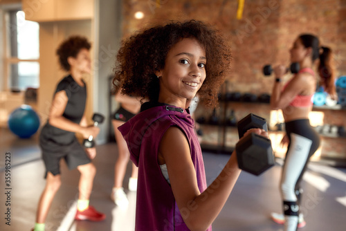 African american girl smiling at camera while exercising using dumbbell in gym together with female trainer and other kids. Sport, healthy lifestyle, physical education concept photo