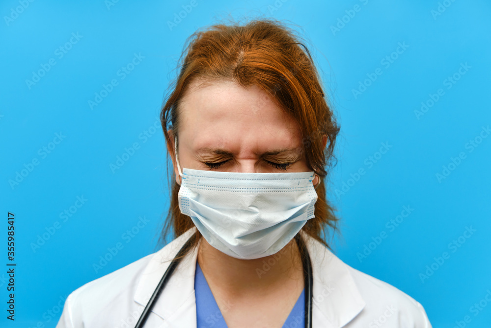 Portrait of a doctor woman with sad facial expression, close up. Nurse in a blue uniform and a medical mask on a blue background.