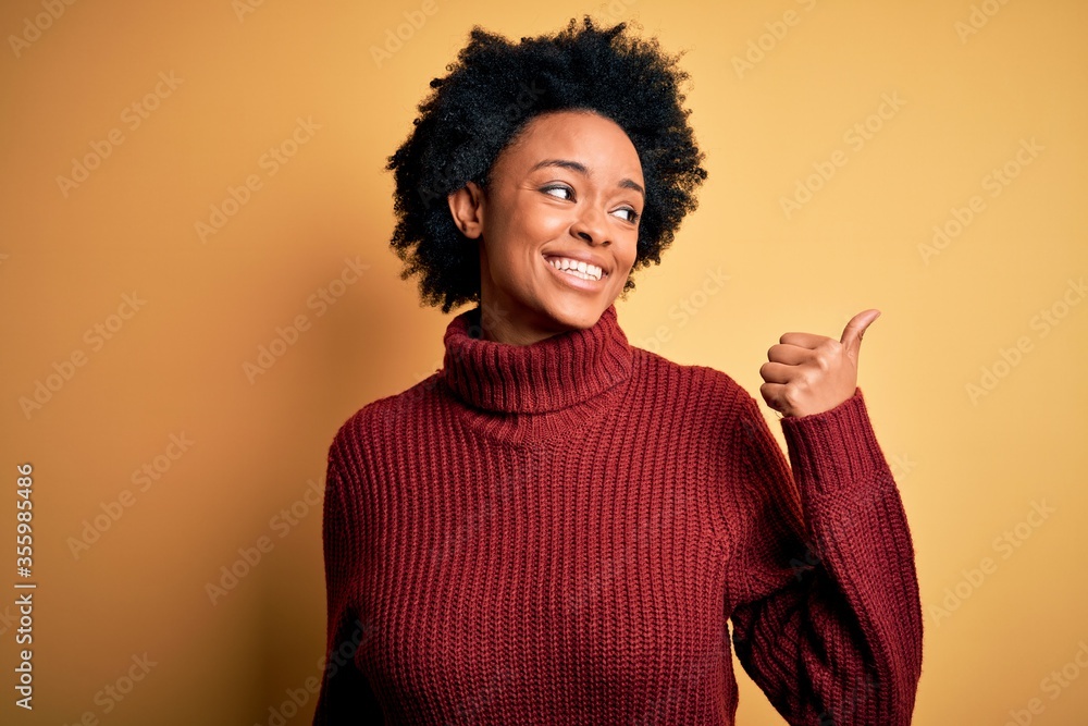 Young beautiful African American afro woman with curly hair wearing casual turtleneck sweater smiling with happy face looking and pointing to the side with thumb up. Young beautiful African American afro woman with curly hair wearing casual turtleneck sweater smiling with happy face looking and pointing to the side with thumb up.