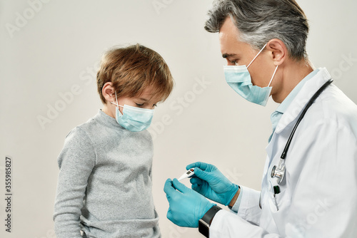 Professional mature doctor pediatrician measuring temperature of a little boy, they are wearing protective masks