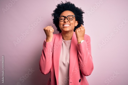 Young beautiful African American afro businesswoman with curly hair wearing pink jacket excited for success with arms raised and eyes closed celebrating victory smiling. Winner concept. photo