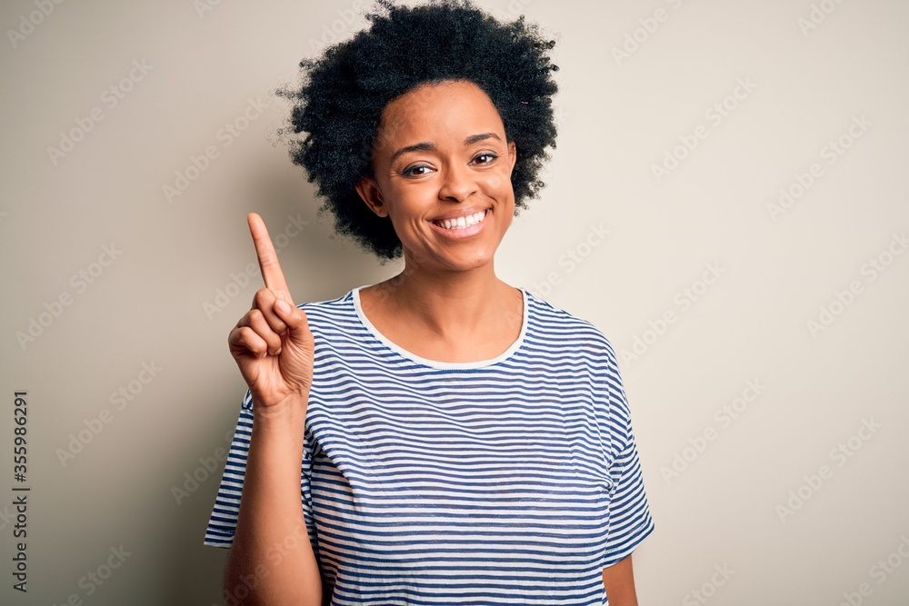 Young beautiful African American afro woman with curly hair wearing striped t-shirt showing and pointing up with finger number one while smiling confident and happy. Young beautiful African American afro woman with curly hair wearing striped t-shirt showing and pointing up with finger number one while smiling confident and happy.