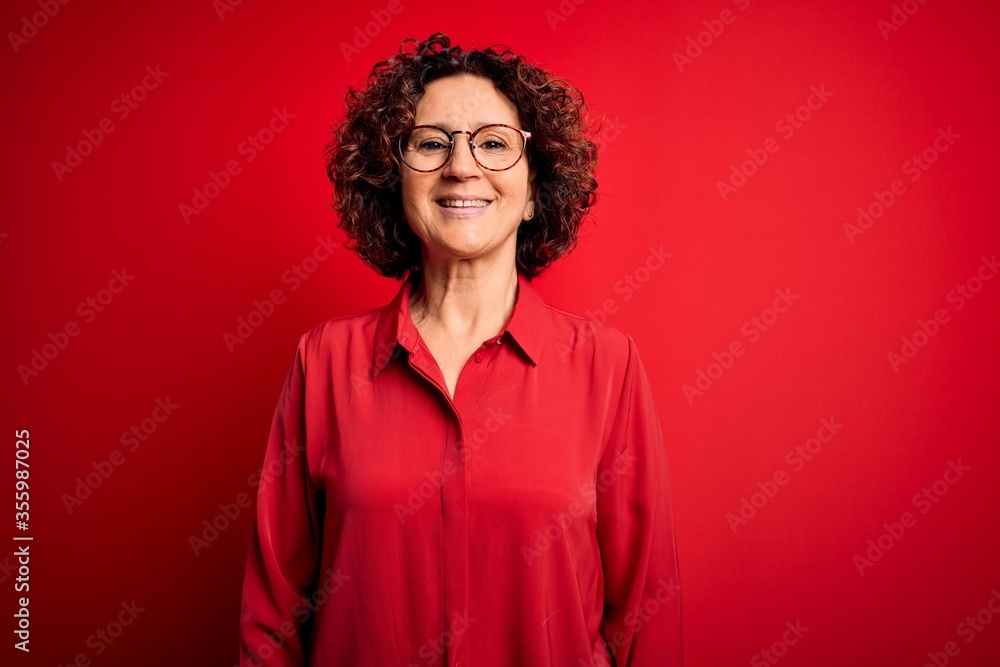 Middle age beautiful curly hair woman wearing casual shirt and glasses over red background with a happy and cool smile on face. Lucky person. Middle age beautiful curly hair woman wearing casual shirt and glasses over red background with a happy and cool smile on face. Lucky person.