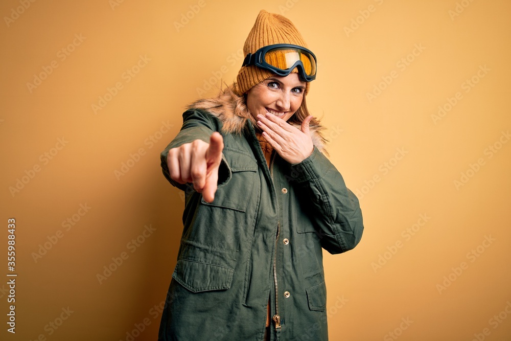 Middle age beautiful blonde skier woman wearing snow sportwear and ski goggles laughing at you, pointing finger to the camera with hand over mouth, shame expression Middle age beautiful blonde skier woman wearing snow sportwear and ski goggles laughing at you, pointing finger to the camera with hand over mouth, shame expression