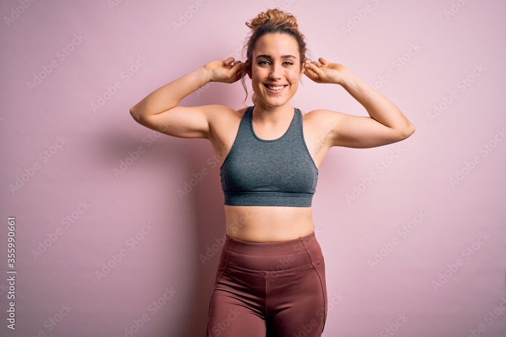 Young beautiful blonde sportswoman doing sport wearing sportswear over pink background Smiling pulling ears with fingers, funny gesture. Audition problem