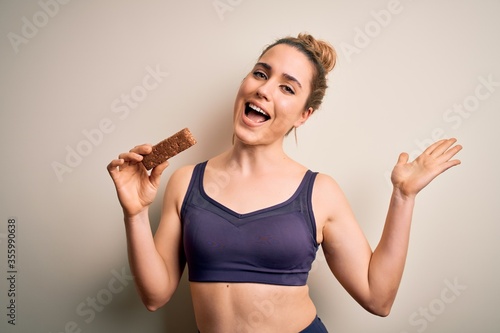Young beautiful blonde sportswoman wearing sportswear holding energetic protein bar very happy and excited, winner expression celebrating victory screaming with big smile and raised hands © Krakenimages.com