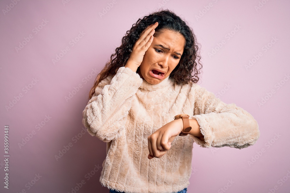 Young beautiful woman with curly hair wearing casual sweater standing over pink background Looking at the watch time worried, afraid of getting late Young beautiful woman with curly hair wearing casual sweater standing over pink background Looking at the watch time worried, afraid of getting late