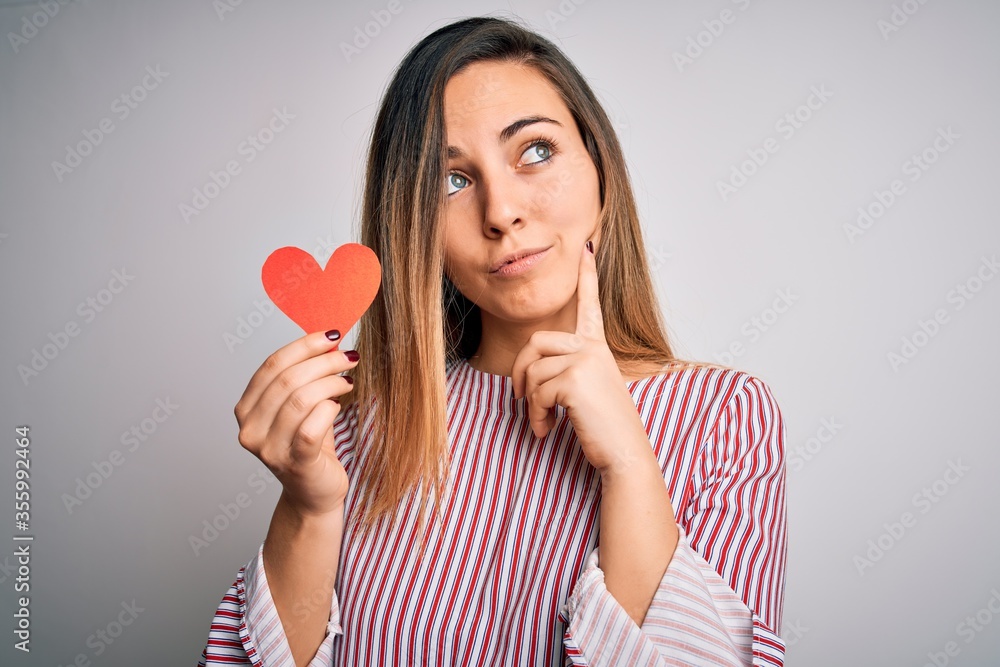 Young beautiful blonde romantic woman holding red paper heart over white background serious face thinking about question, very confused idea Young beautiful blonde romantic woman holding red paper heart over white background serious face thinking about question, very confused idea