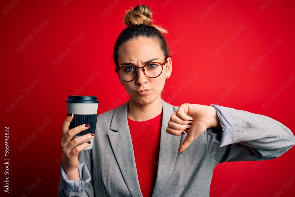 Young beautiful blonde businesswoman with blue eyes wearing glasses drinking cup of coffe with angry face, negative sign showing dislike with thumbs down, rejection concept Young beautiful blonde businesswoman with blue eyes wearing glasses drinking cup of coffe with angry face, negative sign showing dislike with thumbs down, rejection concept