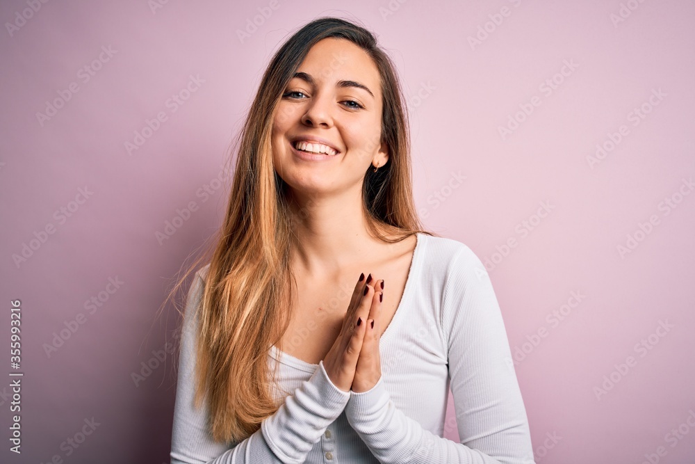 Young beautiful blonde woman with blue eyes wearing white t-shirt over pink background praying with hands together asking for forgiveness smiling confident. Young beautiful blonde woman with blue eyes wearing white t-shirt over pink background praying with hands together asking for forgiveness smiling confident.