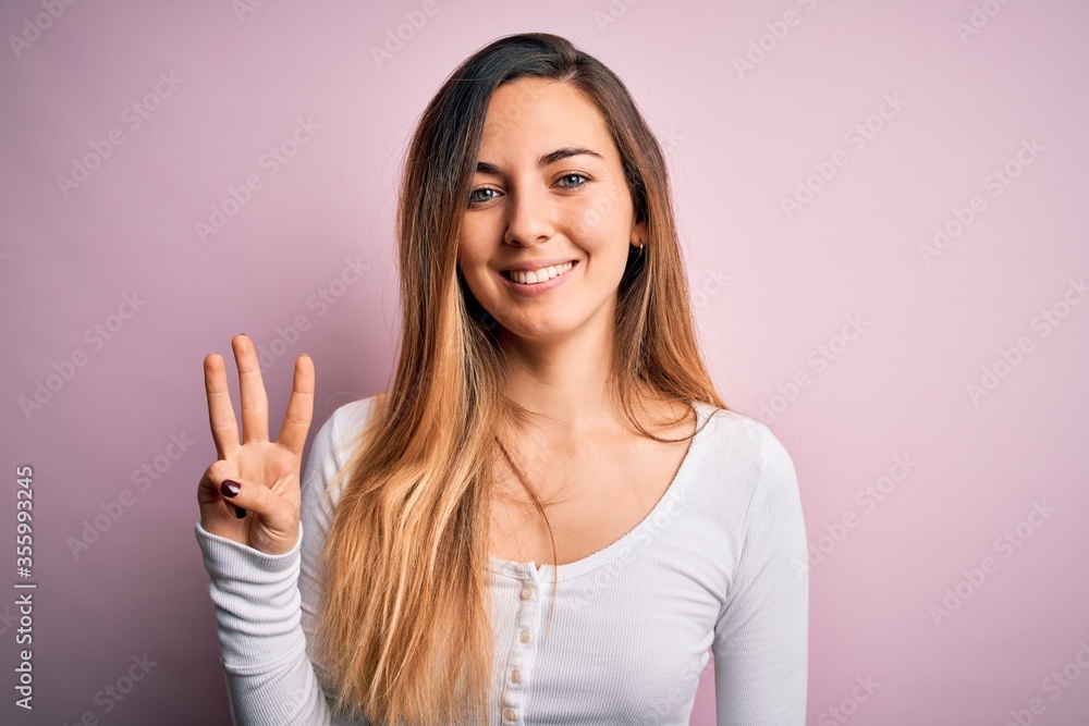 Young beautiful blonde woman with blue eyes wearing white t-shirt over pink background showing and pointing up with fingers number three while smiling confident and happy. Young beautiful blonde woman with blue eyes wearing white t-shirt over pink background showing and pointing up with fingers number three while smiling confident and happy.