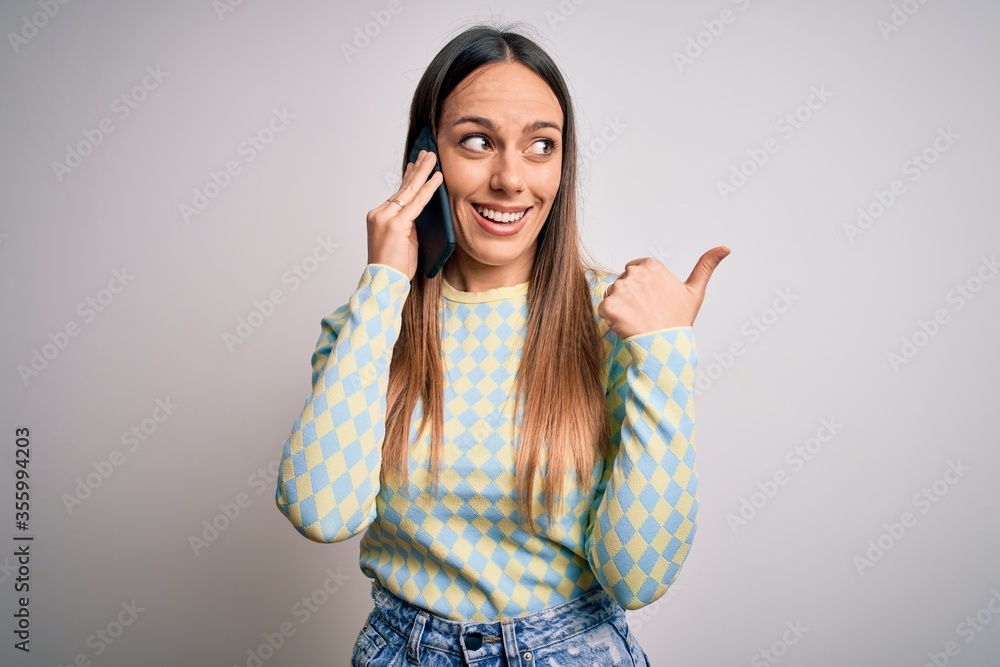 Young blonde woman having a conversation talking on smartphone over isolated background pointing and showing with thumb up to the side with happy face smiling Young blonde woman having a conversation talking on smartphone over isolated background pointing and showing with thumb up to the side with happy face smiling