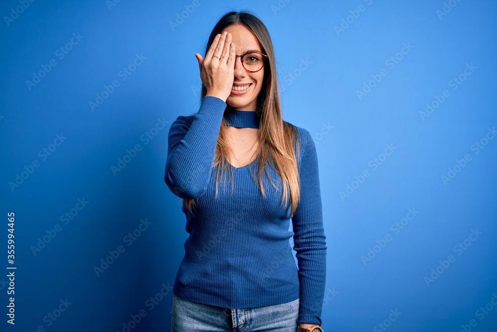 Young beautiful blonde woman with blue eyes wearing glasses standing over blue background covering one eye with hand, confident smile on face and surprise emotion. Young beautiful blonde woman with blue eyes wearing glasses standing over blue background covering one eye with hand, confident smile on face and surprise emotion.