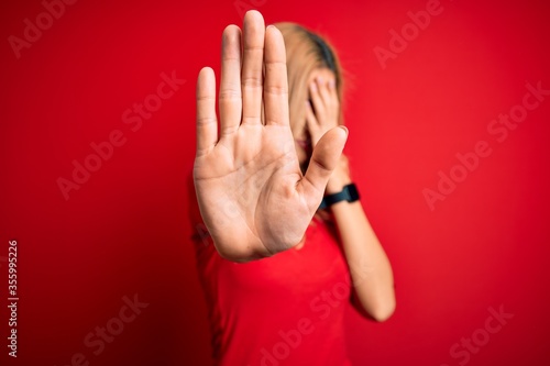 Young beautiful blonde woman wearing casual t-shirt standing over isolated red background covering eyes with hands and doing stop gesture with sad and fear expression. Embarrassed and negative
