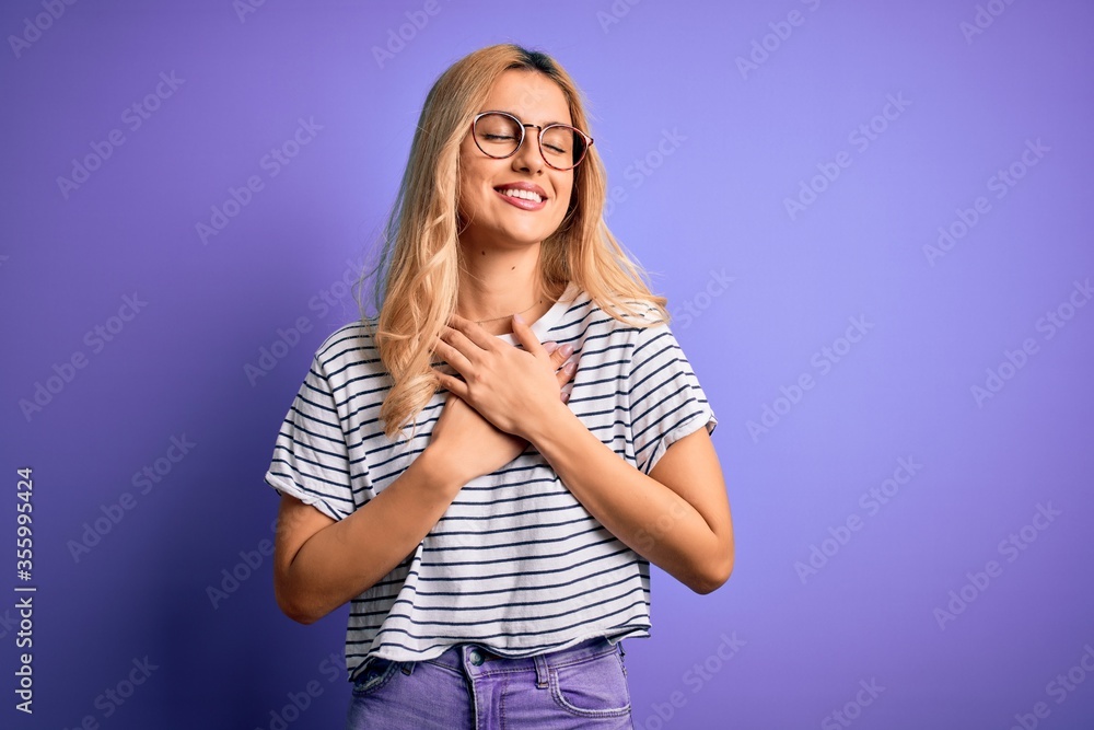 Young beautiful blonde woman wearing striped t-shirt and glasses over purple background smiling with hands on chest with closed eyes and grateful gesture on face. Health concept. Young beautiful blonde woman wearing striped t-shirt and glasses over purple background smiling with hands on chest with closed eyes and grateful gesture on face. Health concept.