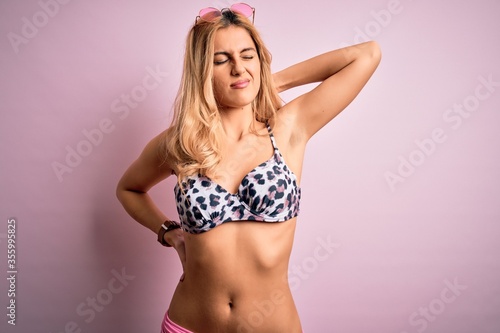 Young beautiful blonde woman on vacation wearing bikini over isolated pink background Suffering of neck ache injury, touching neck with hand, muscular pain
