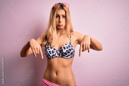 Young beautiful blonde woman on vacation wearing bikini over isolated pink background Pointing down looking sad and upset, indicating direction with fingers, unhappy and depressed.