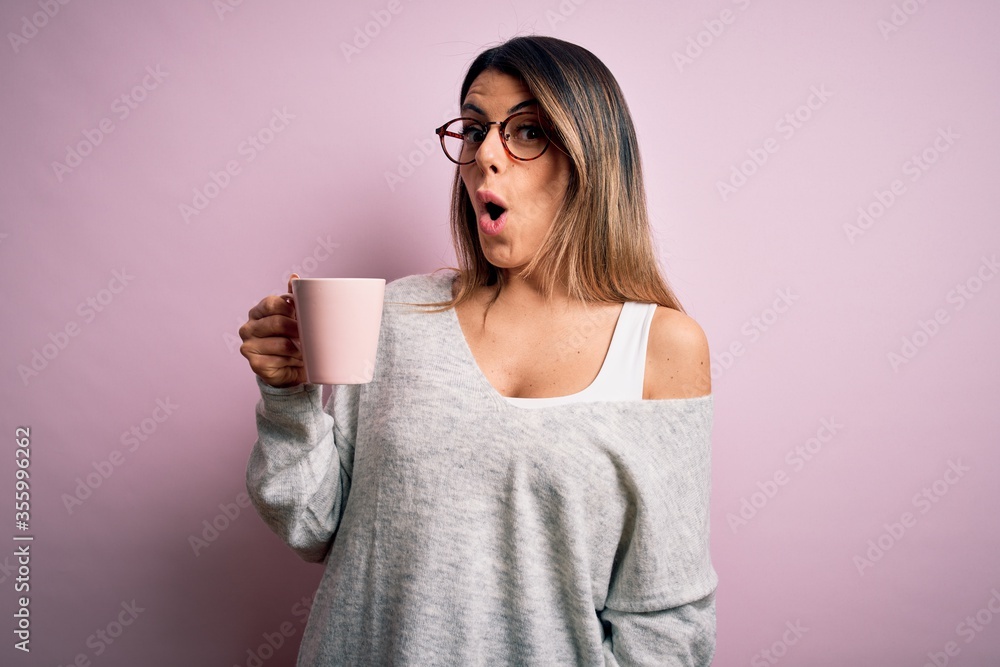 Young beautiful brunette woman wearing glasses drinking pink mug of coffee scared in shock with a surprise face, afraid and excited with fear expression Young beautiful brunette woman wearing glasses drinking pink mug of coffee scared in shock with a surprise face, afraid and excited with fear expression