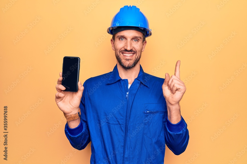 Young handsome worker man wearing uniform and hardhat holding smartphone showing screen smiling happy pointing with hand and finger to the side