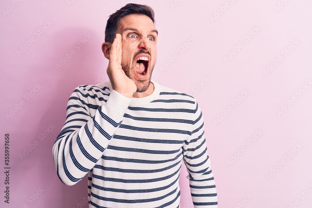 Fototapeta premium Young handsome man wearing casual striped sweater standing over isolated pink background shouting and screaming loud to side with hand on mouth. Communication concept.