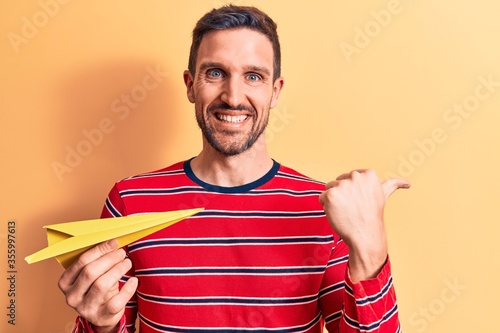 Young handsome man holding yellow paper airplane standing over isolated backgrournd pointing thumb up to the side smiling happy with open mouth photo