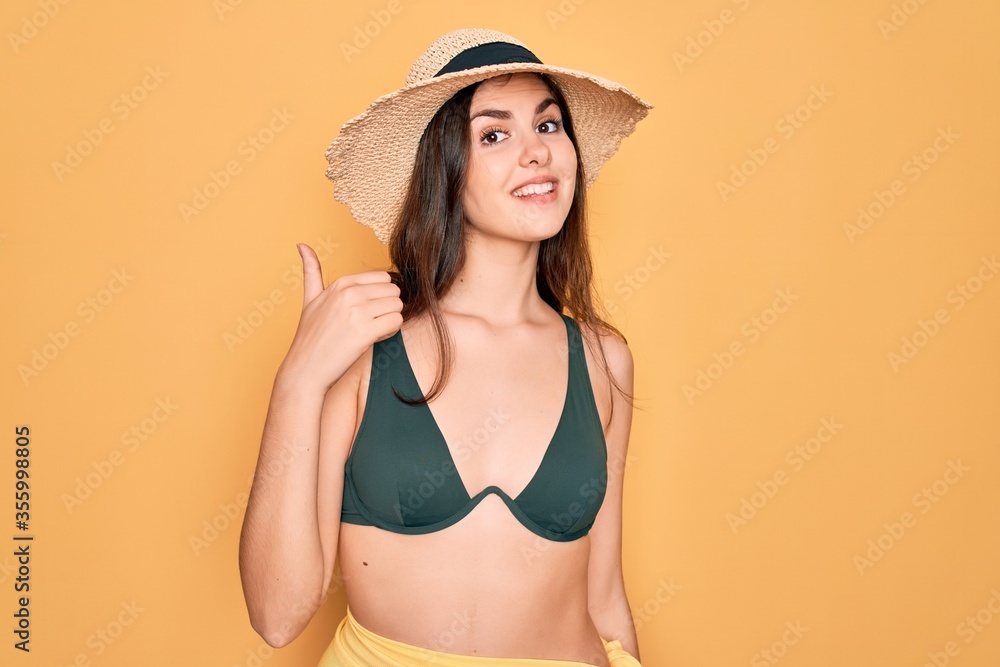 Young beautiful girl wearing swimwear bikini and summer sun hat over yellow background smiling with happy face looking and pointing to the side with thumb up. Young beautiful girl wearing swimwear bikini and summer sun hat over yellow background smiling with happy face looking and pointing to the side with thumb up.