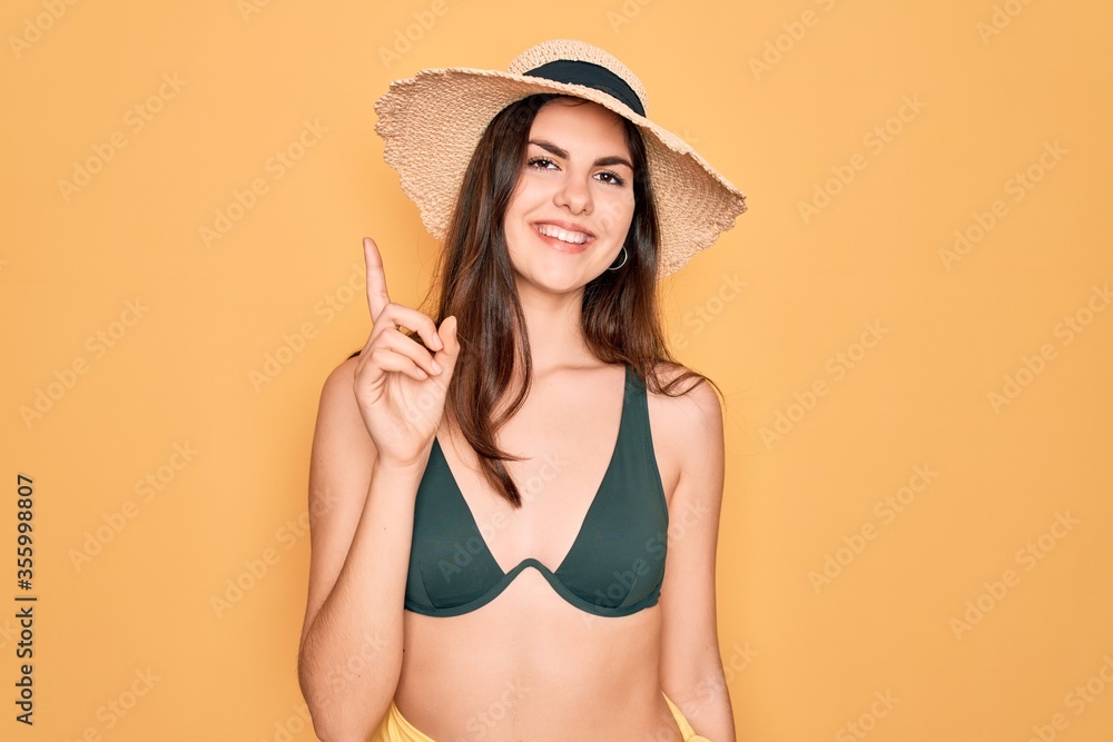 Young beautiful girl wearing swimwear bikini and summer sun hat over yellow background showing and pointing up with finger number one while smiling confident and happy. Young beautiful girl wearing swimwear bikini and summer sun hat over yellow background showing and pointing up with finger number one while smiling confident and happy.
