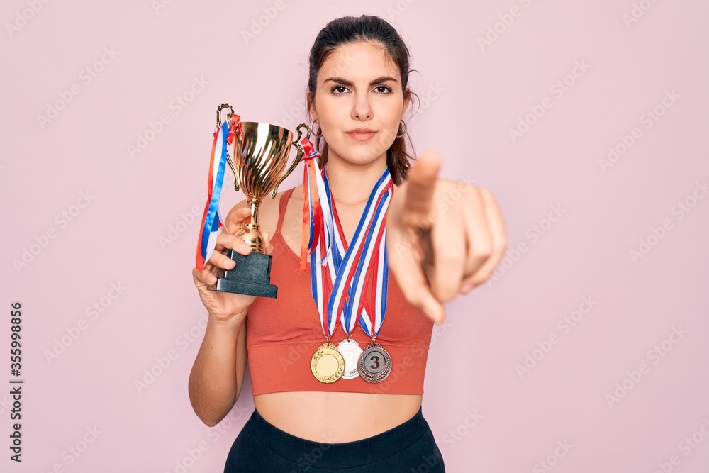 Young beautiful fitness winner athlete woman wearing sport medals and trophy pointing with finger to the camera and to you, hand sign, positive and confident gesture from the front Young beautiful fitness winner athlete woman wearing sport medals and trophy pointing with finger to the camera and to you, hand sign, positive and confident gesture from the front