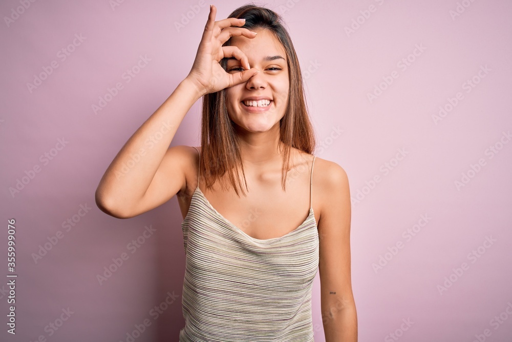 Young beautiful brunette girl wearing casual striped t-shirt over isolated pink background doing ok gesture with hand smiling, eye looking through fingers with happy face. Young beautiful brunette girl wearing casual striped t-shirt over isolated pink background doing ok gesture with hand smiling, eye looking through fingers with happy face.