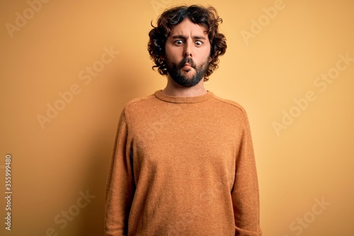 Young handsome man with beard wearing casual sweater standing over yellow background making fish face with lips, crazy and comical gesture. Funny expression.