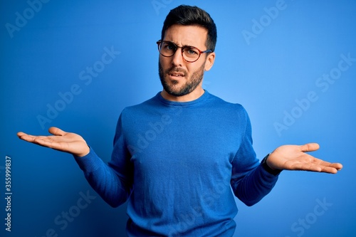 Young handsome man with beard wearing casual sweater and glasses over blue background clueless and confused with open arms, no idea concept. photo