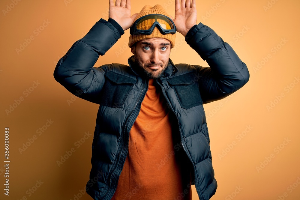 Young handsome skier man with beard wearing snow sportswear and ski goggles Doing bunny ears gesture with hands palms looking cynical and skeptical. Easter rabbit concept. Young handsome skier man with beard wearing snow sportswear and ski goggles Doing bunny ears gesture with hands palms looking cynical and skeptical. Easter rabbit concept.