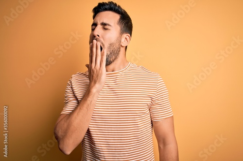 Young handsome man with beard wearing casual striped t-shirt over yellow background bored yawning tired covering mouth with hand. Restless and sleepiness.