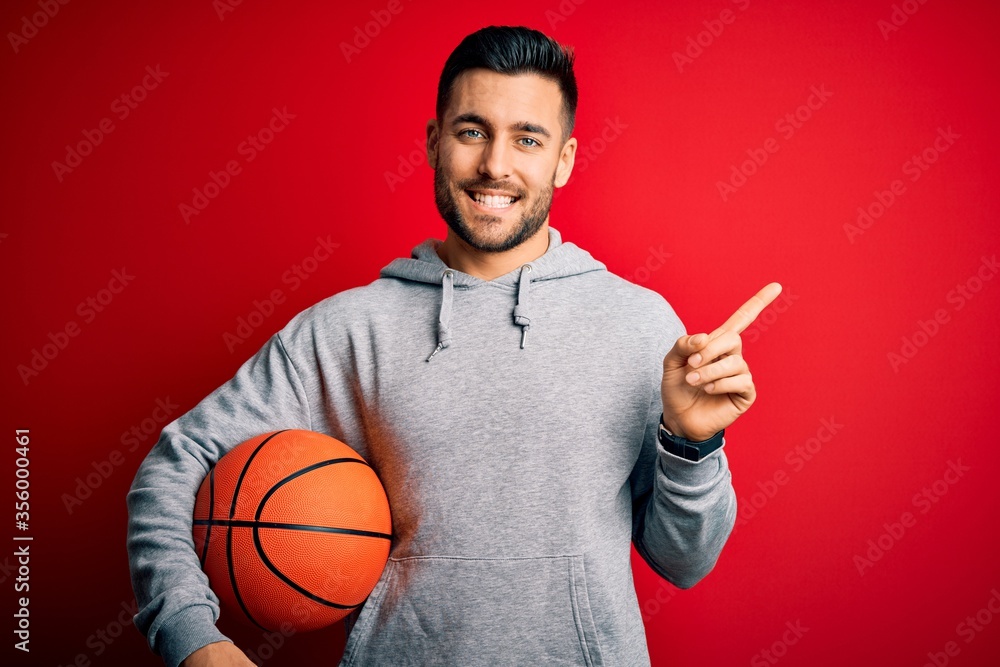 Young sports man holding basketball ball over red isolated background very happy pointing with hand and finger to the side Young sports man holding basketball ball over red isolated background very happy pointing with hand and finger to the side