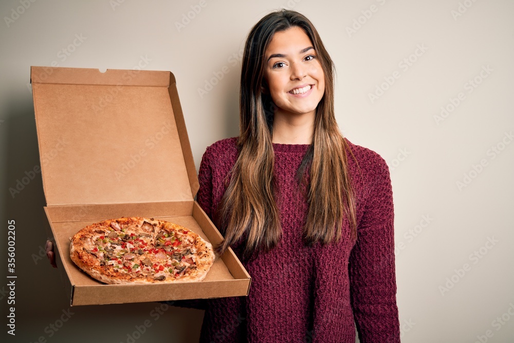 Young beautiful girl holding delivery box with Italian pizza standing over white background with a happy face standing and smiling with a confident smile showing teeth Young beautiful girl holding delivery box with Italian pizza standing over white background with a happy face standing and smiling with a confident smile showing teeth
