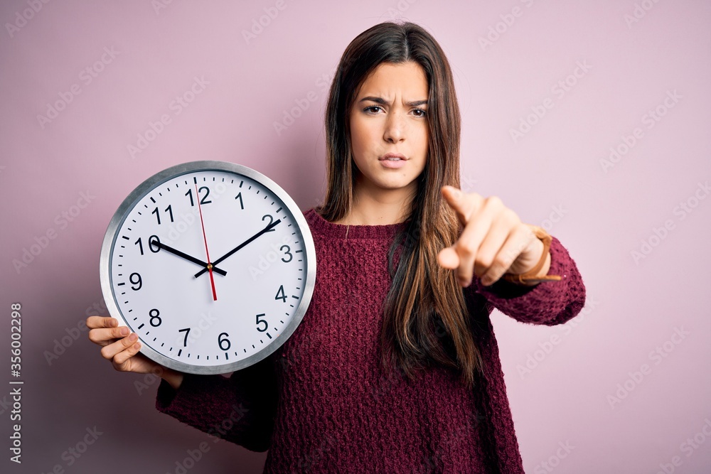Young beautiful girl doing countdown holding big clock over isolated pink background pointing with finger to the camera and to you, hand sign, positive and confident gesture from the front Young beautiful girl doing countdown holding big clock over isolated pink background pointing with finger to the camera and to you, hand sign, positive and confident gesture from the front