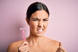 Young beautiful girl using shaver for depilation standing over isolated pink background annoyed and frustrated shouting with anger, crazy and yelling with raised hand, anger concept