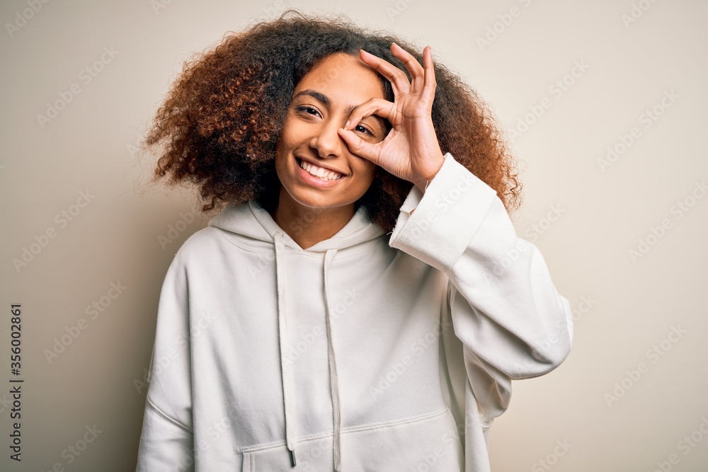 Young african american sportswoman with afro hair wearing sporty sweatshirt doing ok gesture with hand smiling, eye looking through fingers with happy face. Young african american sportswoman with afro hair wearing sporty sweatshirt doing ok gesture with hand smiling, eye looking through fingers with happy face.