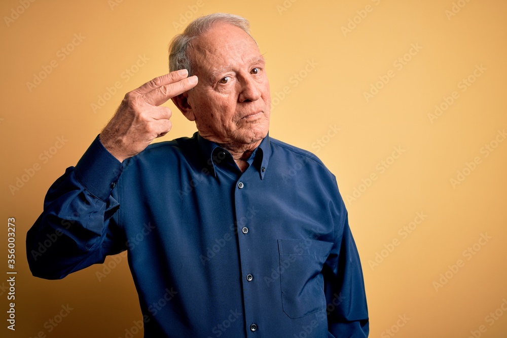 Obraz premium Grey haired senior man wearing casual blue shirt standing over yellow background Shooting and killing oneself pointing hand and fingers to head like gun, suicide gesture.