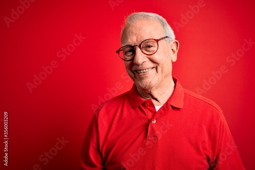 Grey haired senior man wearing glasses and casual t-shirt over red background Relaxed with serious expression on face. Simple and natural looking at the camera. © Krakenimages.com