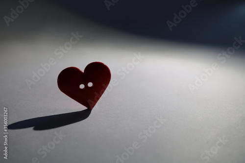 Red love heart button, almost silhouetted by soft light. Casting a long shadow. Lost love, lonely valentine, or bereavement concept. Loss of a loved one. Intentionally underexposed for mood. photo