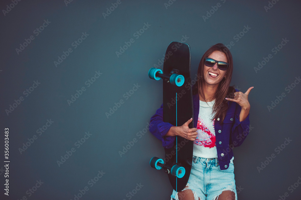 Young happy girl in sunglasses showing shaka with skateboard in front of grey wall. - obrazy, fototapety, plakaty 
