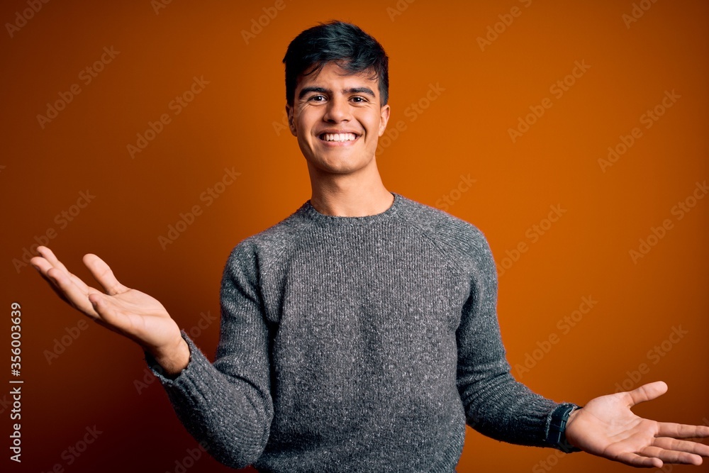 Young handsome man wearing casual sweater standing over isolated orange background smiling cheerful with open arms as friendly welcome, positive and confident greetings Young handsome man wearing casual sweater standing over isolated orange background smiling cheerful with open arms as friendly welcome, positive and confident greetings
